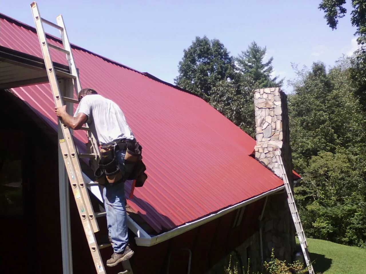 Roofer on a ladder beside a red metal roof and stone chimney — Soffit & Fascia Repair by Roofing Brothers in Kintnersville